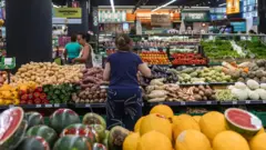 Uma mulherbbb betanocostas escolhendo legumes na seçãobbb betanovegetaisbbb betanoum supermercado com outras pessoas passando ao fundo