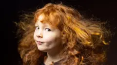 Retrato en primer plano de una niña pequeña con cabello largo, rizado y rojo que ondea como si lo acariciara la brisa. 