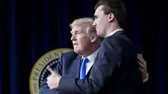 Trump and Kirk embracing in front of a Presidential seal. Kirk is giving a thumbs up to an off-frame crowd