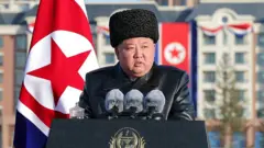 Kim Jong-un habla desde un atril con tres micros. Lleva un gorro y tiene la bandera de su país detrás. 