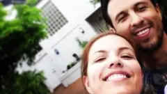Jossette Rivera e seu marido Juan Fernando sorriem numa selfie. O rosto de ambos está no canto direito e há uma construção branca ao fundo