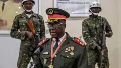 ምሉእ መዓርጉ ዝለበሰ ጀነራል ሆርታ ን'ታም ኣብ ቅድሚኡ ኣብ ዘሎ ማይክሮፎን ቀሪቡ ቃለማሕላ ኣብ ዝፈጸመሉ፡ ክልተ ብረቶም ዘነጻጸሩ ወተሃደራት ብድሕሪኡ ደው ኢሎም ይርኣዩ።