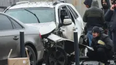 Чоловік у куртці та шапці присів біля пошкодженої вибухом білої машини