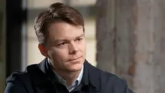 Palantir UK boss Louis Mosley speaking to the BBC. He is wearing a dark jacket with a button up jumper and is sitting in front of a grey wall.