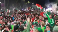 メキシコ代表の緑色のサッカーユニフォームを着た大勢のファンが夜の街で、国旗を振ったり手を上げたりして喜んでいる。大人に肩車した子供も国旗を振っている。紙吹雪のようなものが大勢の頭に積もっている