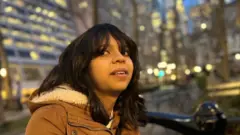 A menina Juliana Peralta, de cabelo escuro, vestindo jaqueta marrom com capuz estofado, fotografada à noite com a visão borrada da cidade atrás dela