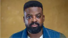 Aworan oṣere tiata, Kunle Afolayan. O wọ aṣọ alawọ aro pẹlu awọtẹnu funfun. Irun ori ati ti ààgbọ̀n rẹ pọ ninu aworan naa, bẹẹ lo n wo ọọkan.