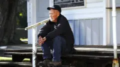 Charlie Hopkins sentado na entrada de sua casa de madeira na Flórida
