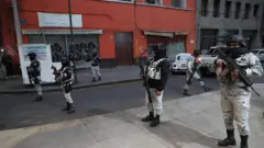 Seis guardas com uniforme cinza-claro, capacetes pretos e lenços pretos cobrindo o rosto fazem a guarda em uma calçada e rua. Eles estão armados com metralhadoras. Um prédio de tijolos vermelhos com janelas fechadas está atrás deles.