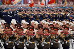 Soldados norcoreanos desfilan en filas portando armas. Los soldados de las primeras filas visten uniformes de color caqui con galones dorados y rojos. Más atrás se encuentran otros soldados norcoreanos con uniformes blancos con galones dorados y negros. Detrás de ellos hay filas de soldados rusos.