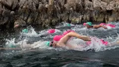 Pessoas com coletes e óculos de natação nadando. Há boias rosas ao lado.