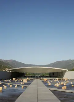 Pasarela de hormigón que conduce a un edificio de techo curvo, rodeado de un estanque con numerosas rocas y montañas al fondo bajo un cielo despejado.