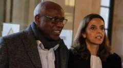 Ibubanfu, Munyemana aboneka yambaye amarori, ishati yera ipfunguye mw'izosi n'igisikafu munsi y'ikoti y'imbeho yirabura, iruhande yiwe haboneka umugore mu mwambaro w'abashingwamanza; bompi bakeye mu maso. 