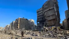 A imagem mostra uma rua de Beirute cercada por prédios danificados, com uma grande estrutura parcialmente destruída e montes de escombros espalhados pelo chão. Algumas pessoas caminham entre os destroços enquanto edifícios ao redor exibem sinais de colapso.