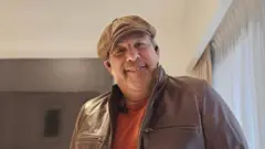 Roberto Guido, um homem de meia idade, vestindo um boné de couro e jaqueta, ambos na cor marrom, com uma camiseta laranja por baixo. Ele sorri para a câmera e tem uma barba rala grisalha