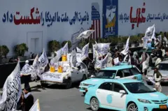 کاروان موترها در یکی از جادههای کابل که در آن پرچمهای طالبان و بشکههای زرد رنگ دیده میشوند.