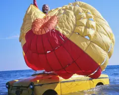 Giorgio Amoretti está de pie sobre un coche amarillo que flota en el aire, sujetando un paracaídas.
