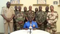Coronel sentado e falando em microfone, rodeado por mais 9 militares em pé