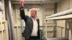 Janusz, de jeans e jaqueta xadrez, aponta para o teto de um de seus abrigos. O local tem um beliche e um vaso sanitário com cortina. É limpo, mas espartano.