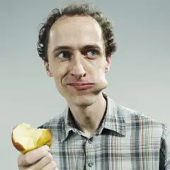 Homem comendo maçã e olhando para o lado, com um pedaço da fruta na boca