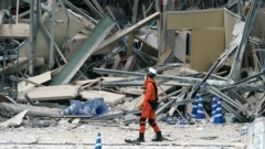 Un bombero, con uniforme anaranjado, botas negras, casco y mascarilla blancos, pasa por el lado de los escombros de una infraestructura 