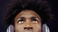 Jovem negro com cabelo afro olha para cima; ele usa fones de ouvido tipo headphones