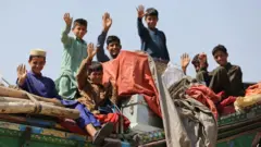 شش کودک افغان روی اثاثیه‌های بار کامیون نشسته‌اند و با لبخند دست تکان می‌دهند