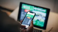 Celular com aplicativo de apostas, e tela atrás com aparentemente um jogo esportivo