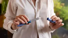 Zdjęcie stockowe przedstawia zbliżenie osoby trzymającej w dłoniach dwie części zastrzyku na odchudzanie. Zastrzyk wygląda jak długopis z zdjętą nasadką, a osoba ma na sobie różową bluzkę.