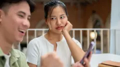 Umugore utishimye ukomoka muri Aziya yafushye, bitewe n'uburyo umukunzi we ahugiye kuri telefone yishimye asa nk'uwamuburiye umwanya. 