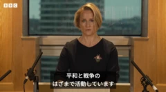 カメラに向かって演説する女性。金髪ショートボブで、暗い色のカットソーを着ている。胸元には甲虫のブローチを着けている