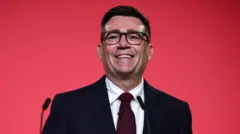 赤い背景のステージで笑顔で演説するバーナム氏。黒いスーツにえんじ色のネクタイを着けている