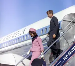 Jackie Kennedy descendo as escadas de um avião. Ela veste um conjunto de saia e blazer rosa, com chapéu rosa. Atrás dela está o marido, Jhon Kennedy, de terno.