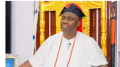 Alaafin Oyo, Oba Abimbola Owoade, jokoo lori itẹ, o de fila abetiaja alawọ aro. 
O wọ aṣọ funfun pẹlu ilẹkẹ pupa to fi sọrun.
