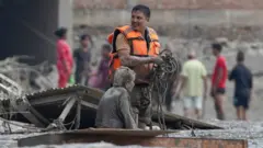 Un hombre con un chaleco refectante naranja está de pie en un espacio al aire libre junto a una mujer sentada en el piso y cubierta de lodo.