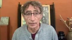 Um homem vestindo um suéter cinza e óculos fala para a câmera.
