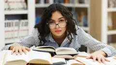 Mujer joven estudiando con rostro de frustración junto a varios libros abiertos