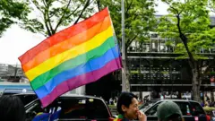 男性が性的マイノリティー（LGBTQ）の権利を象徴する大きなレインボーフラッグを掲げて、大通りを歩いている。旗は大きく風にはためいている。車道にはタクシーが停車しており、反対側の車線や歩道、建物なども見える