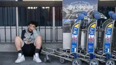 Un hombre sentado en el piso junto a varios carritos para transportar maletas típicas de los aeropuertos. El hombre tiene la mano sobre el rostro con un gesto que denota agotamiento.