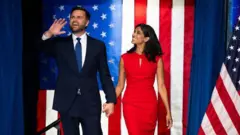 JD Vance ve eşi Usha Vance ABD bayrağı fonunda sahneye çıkarken el sallıyor