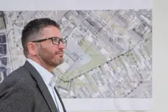 Un hombre con chaqueta gris, camisa clara y gafas mira hacia un lado. Tiene el pelo corto y oscuro con canas. Está de pie frente a un mapa topográfico superpuesto a la imagen de un yacimiento.
