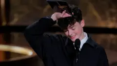 Owen Cooper vestido con traje oscuro y camisa blanca, sonríe y mira con rubor hacia el piso, mientras coloca su mano derecha sobre su cabeza.