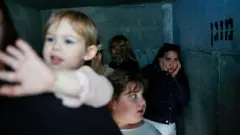 Невелике укриття з бетонними стінами, де перебуває група молодих жінок і дітей. Тісний простір освітлений слабким холодним світлом, одна з дівчат дивиться стривожено в камеру, притискаючи руки до обличчя.