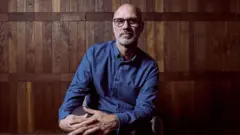 Jesse Armstrong de óculos e camisa azul sentado de pernas cruzadas olhando para a câmera