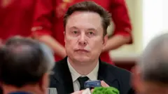 Elon Musk sentado, segurando um iPhone em um evento, vestindo terno e gravata, com uma mulher vestida de vermelho ao fundo