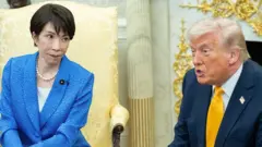 青いジャケットにパールのネックレスを着けた高市氏と、ダークスーツに金色のネクタイを着けたトランプ氏が、クリーム色の布張りの椅子に並んで座っている。高市氏は口を閉じ、記者団に向かって話すトランプ氏を見ている