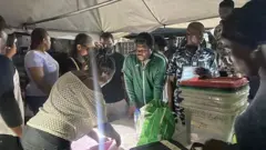 Mr Macaroni àtàwọn ọ̀dọ́ mìíràn tí wọ́n ń dúró láti ri pé àwọn òṣìṣẹ́ fi èsì ìbò ṣọwọ́ sórí IReV