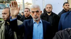 Yahya Sinwar, com um dos braços para o alto, rodeado de integrantes do Hamas. Ele veste uma camisa azul clara e um terno azul. Todos os homens vestem roupas civis.
