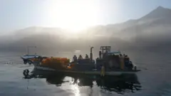 Um pequeno barco de pesca com pessoas a bordo é visto na água, com neblina e montanhas ao fundo.
