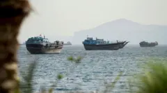 Boats atop the water pictured in a view of the Strait of Hormuz on a hazy day in mid-May with rock and plantation bokeh in the foreground and mountains in the background, taken from Oman on 22 May.